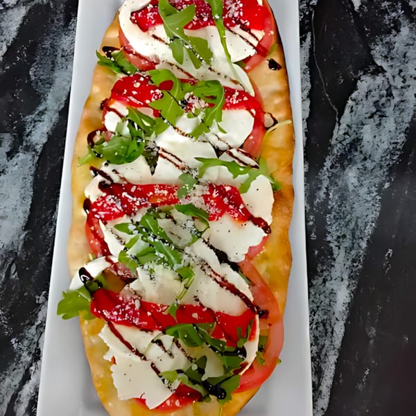 A long flatbread pizza with mozzarella, tomatoes, arugula, and balsamic glaze on a white platter.